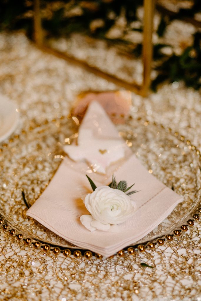 Pink hemstitch napkins adorned with ranunculus blooms and custom pink and white marbled Texas shaped cookies , on glass chargers accented with gold beads.  